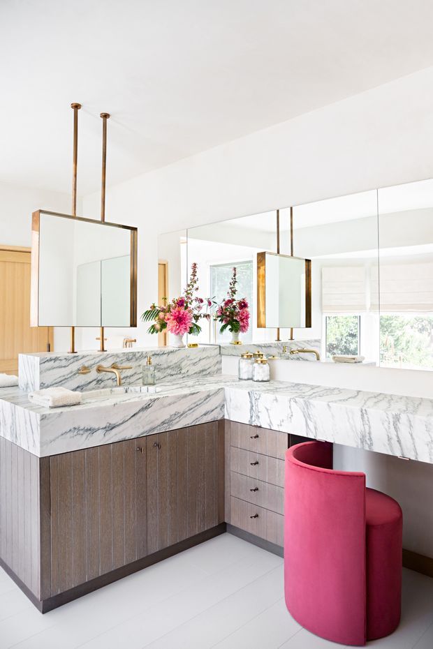 Bright, refined bathroom with a double vanity arranged in an L-shaped configuration. The room features dramatic white-and-grey veined marble countertops and integrated backsplash, dark vertically textured wood cabinetry, large mirrored wall panels, twin suspended rectangular mirrors with bronze-toned frames, brass tapware and hardware, white tiled flooring, and a vivid pink upholstered vanity stool. A floral arrangement adds colour and softness.