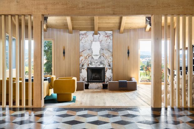 A warm, wood-lined living room framed by vertical timber slats and substantial timber posts. The room features a sculptural fireplace with a dramatic veined stone surround, colourful upholstered seating, a low ottoman, exposed timber ceiling beams, and a highly patterned geometric tile floor in the foreground. Large windows and glazed openings provide views toward a leafy landscape.