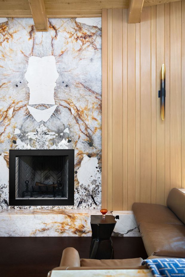 A refined living room centered on a dramatic floor-to-ceiling natural stone fireplace wall with prominent white, grey, and warm gold veining. The room features vertical light timber wall panelling and exposed timber ceiling beams, a black fireplace surround with dark herringbone firebox lining, a dark brown floor, and a tan leather sofa and armchair. A sculptural black side table and slim brass wall light add contrast. The overall composition is warm, luxurious, and strongly material-focused.