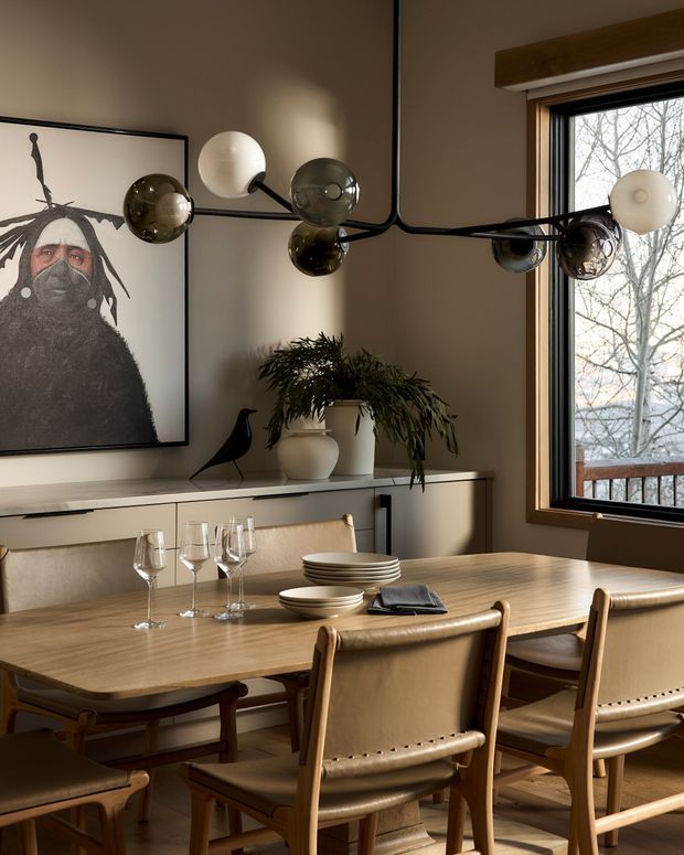 Warm, refined dining room with a substantial light timber dining table and matching leather-and-wood dining chairs. A sculptural black branching pendant with globe lights hangs above the table. The room includes a low built-in sideboard, framed artwork, a potted plant, and a large timber-framed window overlooking trees and a balcony or deck.