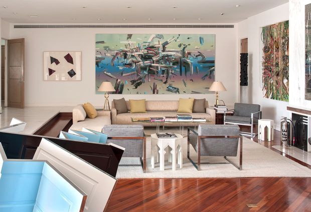 Bright, spacious contemporary living room with a large abstract artwork, beige sectional sofa, upholstered lounge chairs, central glass coffee table, recessed ceiling lighting, polished hardwood flooring, and a large decorative wall installation. A piano or console occupies the foreground, while open doorways and built-in wall features frame the room.