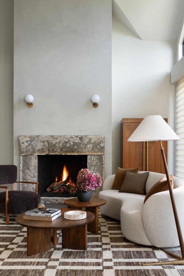 A refined living room with a tall pale plaster fireplace wall and a rough natural-stone hearth surround. The room features a lit fire, sculptural cream upholstered seating, dark timber nesting coffee tables, a mid-century style wooden cabinet, globe wall sconces, a large angled floor lamp, and a geometric brown-and-cream rug. Warm wood, stone, and neutral upholstery create a sophisticated, cosy atmosphere.