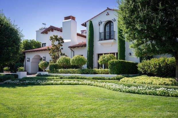 Exterior view of a large white stucco residence with terracotta-tiled roofs, arched openings, wrought-iron balcony railings, decorative wall lanterns, and a prominent chimney. The home is surrounded by meticulously clipped hedges, columnar evergreens, layered garden beds, and a broad manicured lawn under bright sunlight.