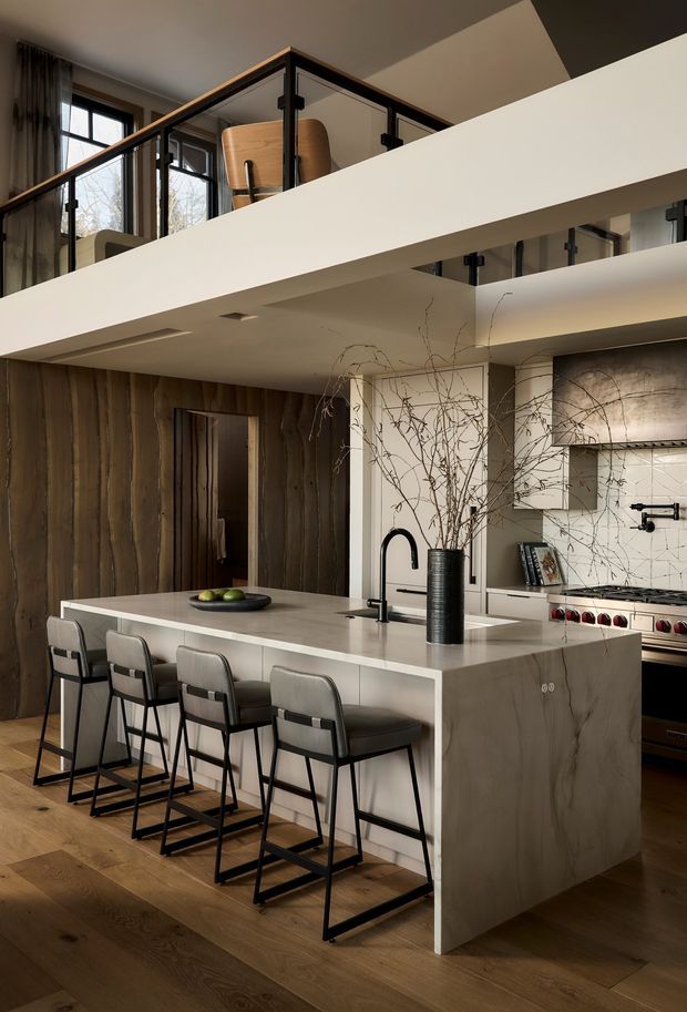 A sophisticated double-height kitchen with a large pale stone island and integrated seating for four. The kitchen features dark timber wall cladding, warm timber flooring, white tiled splashback, dark cabinetry, a black gooseneck tap, and a concealed cooking area. A mezzanine level with black-framed glass balustrades overlooks the kitchen, while exposed branches in a tall vase add an organic decorative element.