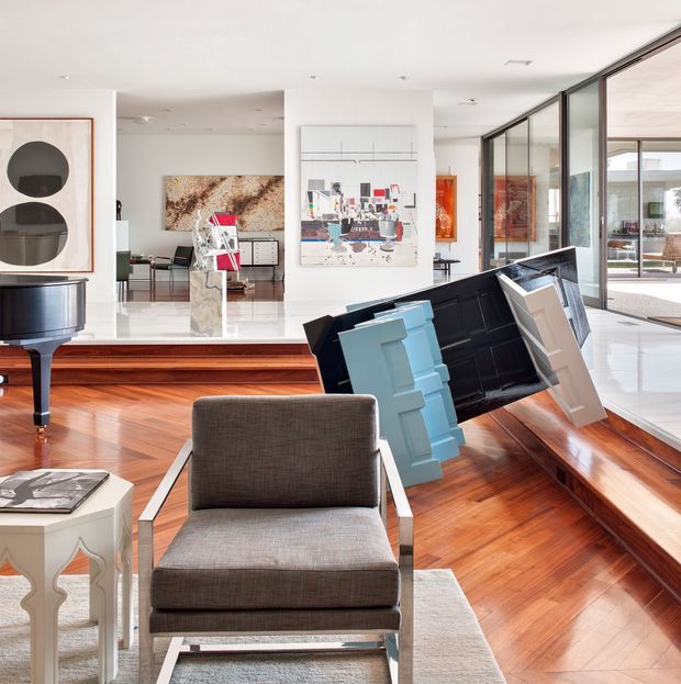 Bright, spacious contemporary living room with polished warm timber flooring, a grey upholstered armchair, a grand piano, abstract artwork, and a sculptural black shelving or console piece with colourful geometric forms. Full-height black-framed glazing lines one side of the room, opening the space visually to an outdoor area. White walls and ceiling, stepped floor levels, and curated art create a refined gallery-like interior.