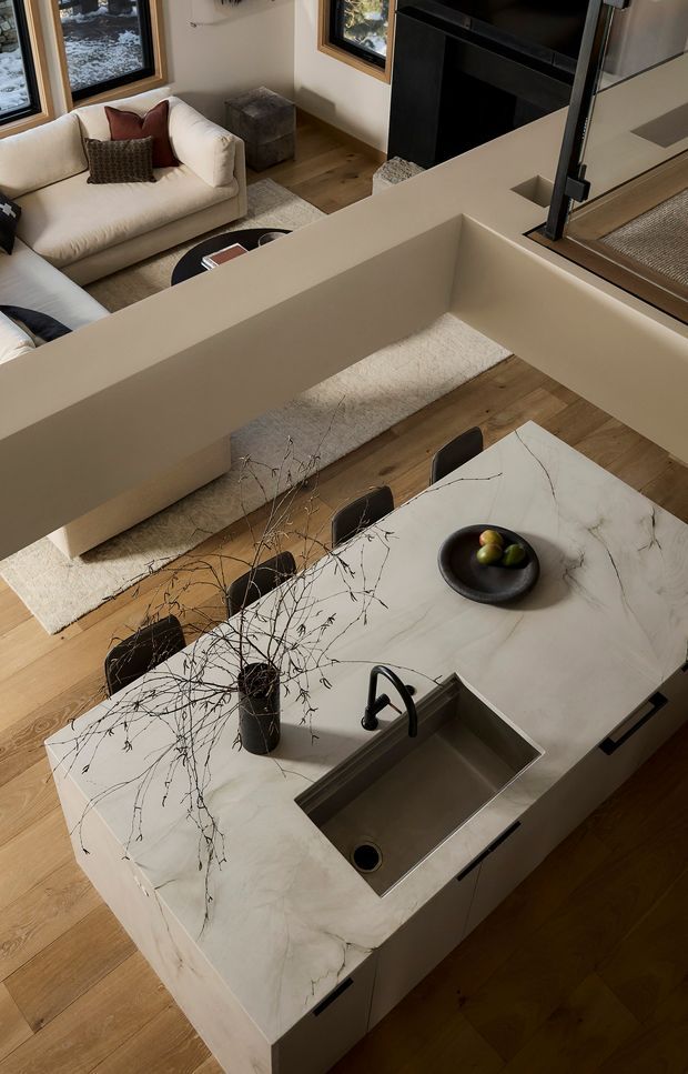 Elevated view of an open-plan kitchen and living area. A large island with a veined light stone benchtop, integrated stainless-steel sink and dark tap dominates the foreground, with dark fixed cabinetry below. Timber flooring extends through the space. Behind the island is a dining table and chairs, a cream sofa with cushions, a rug, windows with timber frames, and a dark fireplace or built-in feature wall. The interior has a warm, refined contemporary aesthetic with neutral finishes and strong connection to the surrounding landscape through the windows.