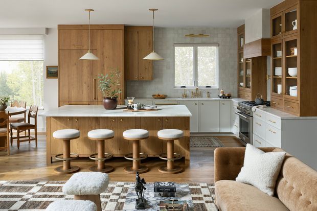 Warm, inviting open-plan kitchen and living area with a large timber island, four upholstered stools, white cabinetry, pale tiled backsplash, integrated appliances, and extensive built-in timber shelving. A dining area is visible to the left and a tan sofa occupies the foreground, with layered rugs and natural timber flooring adding texture.
