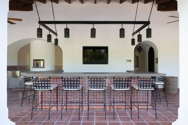 A spacious covered outdoor bar and entertaining area with a long tiled counter, patterned bar stools, dark metal hanging pendant lights, exposed timber roof beams, terracotta floor tiles, white plaster walls, arched openings, and a television. The space has a Mediterranean-inspired architectural character with a strong geometric, patterned interior fit-out.