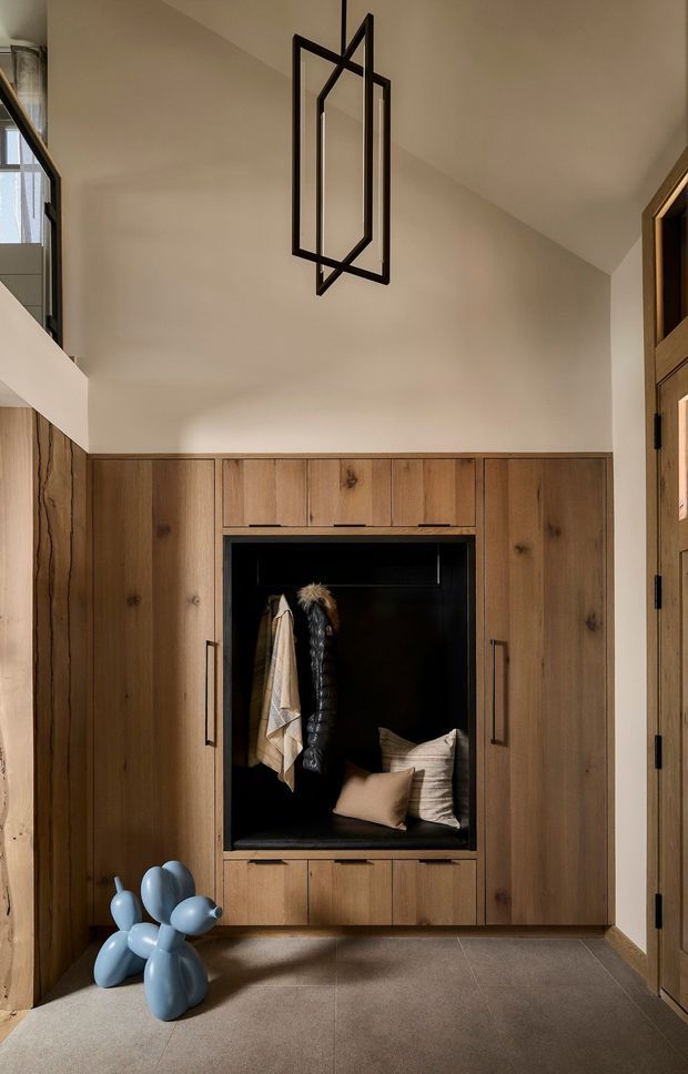 A warm, double-height entryway or hallway featuring extensive built-in timber cabinetry with a central black-lined coat storage nook and bench. The space has pale plaster walls, large grey floor tiles, a sculptural black geometric pendant, and a glimpse of an upper-level landing and window. A blue balloon-dog sculpture adds a playful accent.