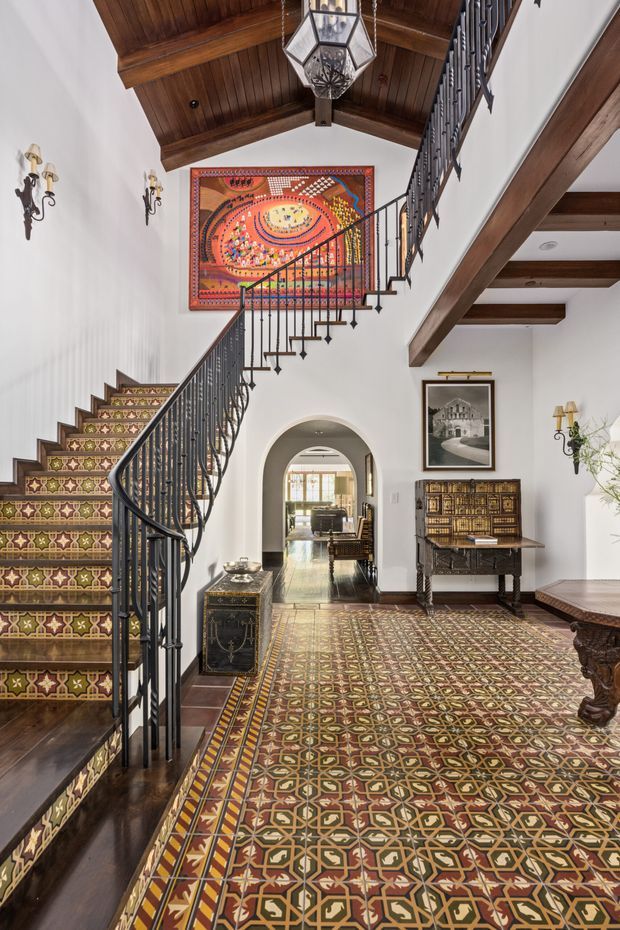 A grand interior stair hall with an ornate patterned tile floor and matching tiled staircase risers. A sweeping staircase has dark wrought-iron balustrades, while white plaster walls, exposed dark timber beams, and a vaulted timber ceiling create a richly detailed Mediterranean-inspired setting. Decorative wall sconces, a large colourful artwork, antique-style furniture, and an arched passageway add to the formal character.