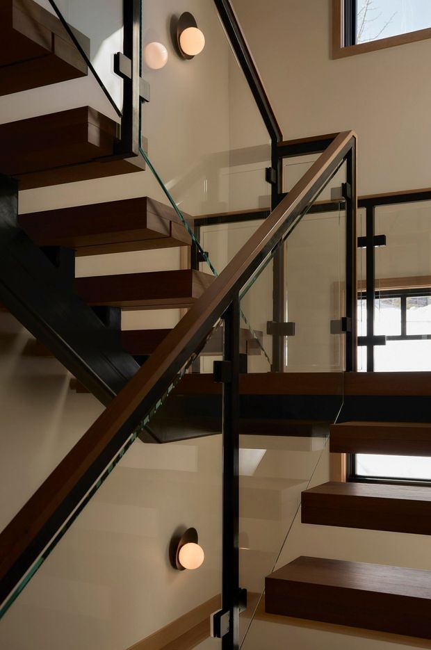 Interior view of a contemporary stairway with floating dark timber treads, black structural supports, and clear glass balustrades. The stair forms a dramatic geometric composition around a central void, with warm wall sconces, pale walls, and timber-framed windows providing natural light.