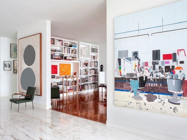 Bright, refined contemporary interior featuring a polished light stone foyer floor, white walls, a large abstract artwork, and a dark green accent chair. An open library area beyond contains extensive built-in white shelving filled with books and artwork, with a glossy dark timber floor. The space has abundant natural light and a curated art-gallery atmosphere.
