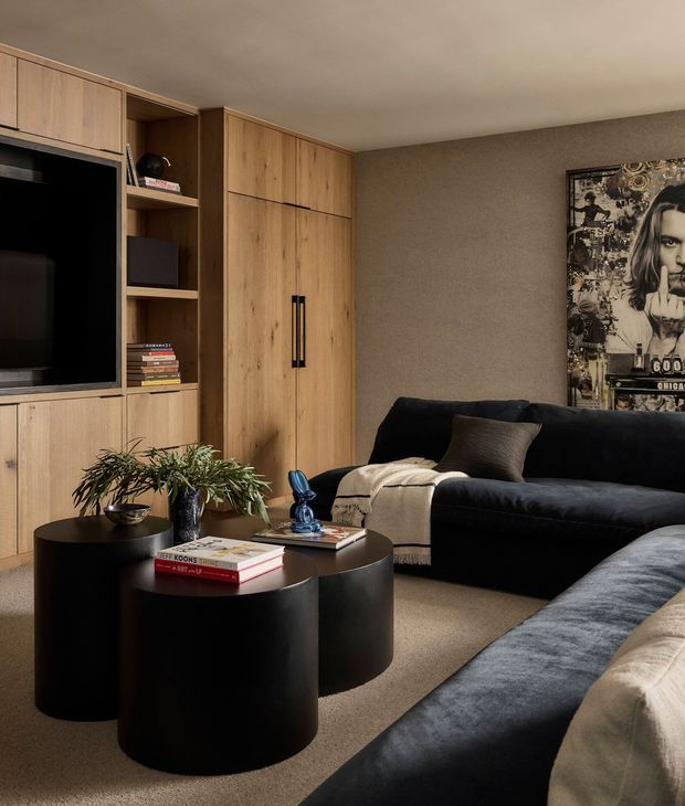 A sophisticated living room with full-height integrated timber cabinetry, a built-in television and open shelving, paired with deep charcoal sofas, black cylindrical nesting coffee tables, a neutral area rug, and a large monochrome artwork. The space has warm, muted tones and a refined contemporary feel.