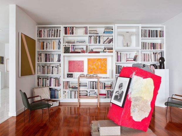 Bright, curated living room or study with extensive floor-to-ceiling white built-in bookshelves filled with books and art objects. The room has polished warm timber flooring, white walls and ceiling, mid-century-style lounge chairs, framed artwork, and a large colourful abstract artwork leaning in the foreground. The space feels artistic, refined, and book-lined, with a compact reading or sitting area.