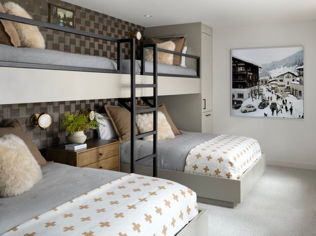 A compact, stylish bunk bedroom with two built-in bunk beds arranged along the walls. The room features grey bedding, patterned throws, textured dark geometric wall panels, integrated shelving and lighting, a central black ladder, a timber bedside cabinet, and a pale carpeted floor. A framed snowy alpine village photograph adds a mountain-inspired accent.