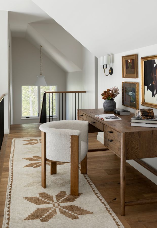 A refined home office or study on an upper-level landing, featuring a substantial mid-tone timber desk with drawers, a softly upholstered armchair, patterned neutral rug, framed artwork, and a contemporary wall sconce. A timber-and-black-metal balustrade overlooks a stairwell beside tall windows, while white walls and ceiling create a bright, calm setting.