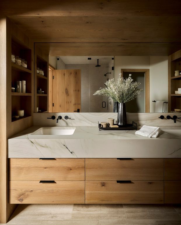 A warmly lit contemporary bathroom vanity with a wide double-basin counter, veined light stone benchtop, timber drawer cabinetry, dark fixtures, large mirror, open timber shelving, and a walk-in shower visible in reflection. The space has a refined natural material palette and integrated storage.