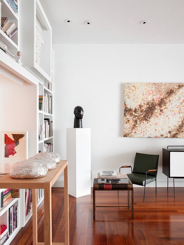 Bright, refined living room or reading space with white built-in floor-to-ceiling shelving filled with books, polished timber flooring, a simple timber table, mid-century-style dark green armchair, coffee table, sculptural bust, and abstract artwork. The room has a clean white palette with warm wood accents and carefully curated contemporary art and furnishings.