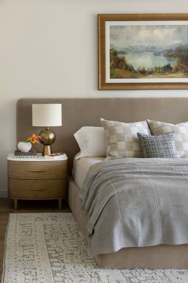 A softly styled bedroom vignette featuring a taupe upholstered bed with layered white and grey bedding, textured accent cushions, a rounded mid-century-inspired timber bedside table, and a brass table lamp. A framed landscape artwork hangs above the bed, with a patterned neutral rug underfoot. The room has a calm, refined, warm-neutral aesthetic.