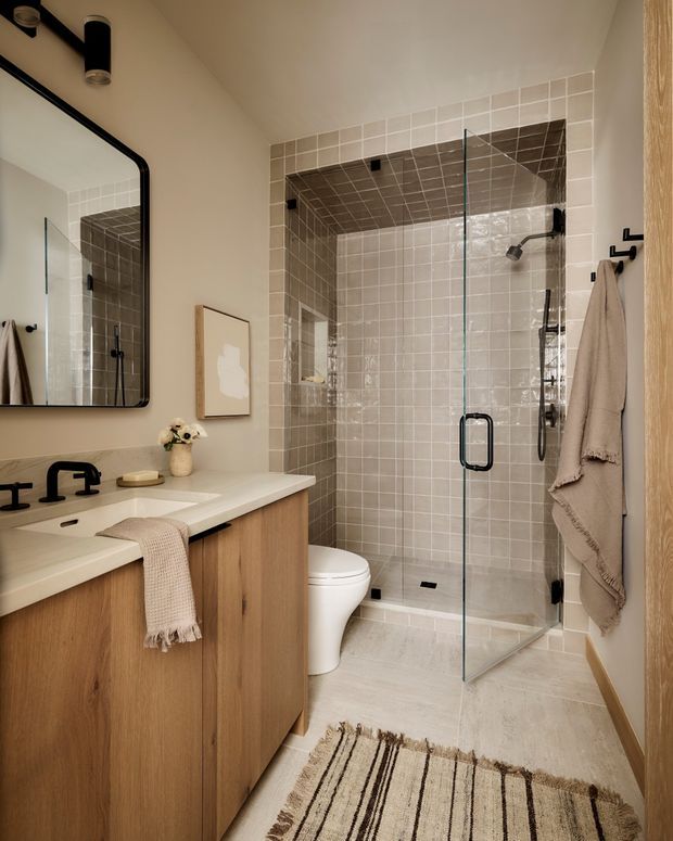 A warm, contemporary bathroom featuring a long timber vanity with a white integrated countertop, matte black tapware and hardware, a large rounded-corner mirror, and a fully tiled walk-in shower enclosed by clear glass. The shower and partial walls use small square beige-grey tiles, while the floor has a light stone-like finish. A toilet, wall-mounted towel hooks, and neutral textiles complete the space.