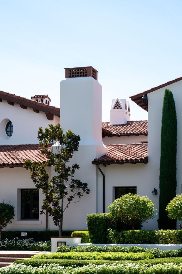 Exterior view of a refined Spanish or Mediterranean-style residence with white stucco walls, red clay tiled pitched roofs, prominent stucco chimneys with decorative brick and lattice caps, dark timber roof brackets, arched and circular windows, and formal manicured hedges and garden beds in the foreground.