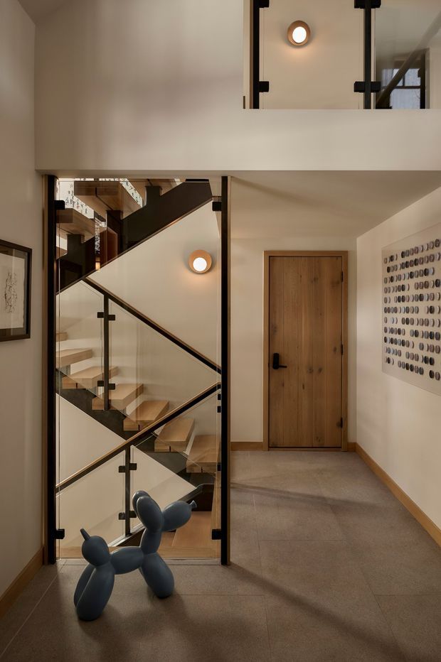 A warm, contemporary residential hallway and stairwell viewed from the entry area. The space features a multi-level staircase enclosed by clear glass panels with dark metal fittings and timber handrails, a natural timber door, large pale floor tiles, warm off-white walls, and minimalist wall sconces. A sculptural black balloon-dog artwork and framed wall art add decorative accents.