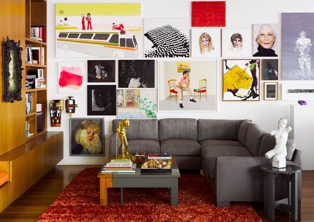 Eclectic living room with a large grey sectional sofa, dark timber built-in shelving and cabinetry, a low coffee table, and a deep red textured rug. The white walls are densely arranged with a varied gallery of paintings, prints, and photographs, creating a colourful art-focused interior. Decorative sculptures and books add further visual interest.
