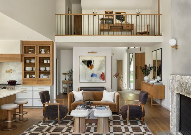 A sophisticated open-plan living space with a visible kitchen at left, a central seating area, and a mezzanine-level study or lounge above. The room features warm timber flooring, white walls, built-in timber shelving and cabinetry, a large abstract artwork, a patterned rug, mid-century-inspired furniture, and a substantial stone fireplace. An open stair or circulation area connects to a glazed exterior door at the rear.
