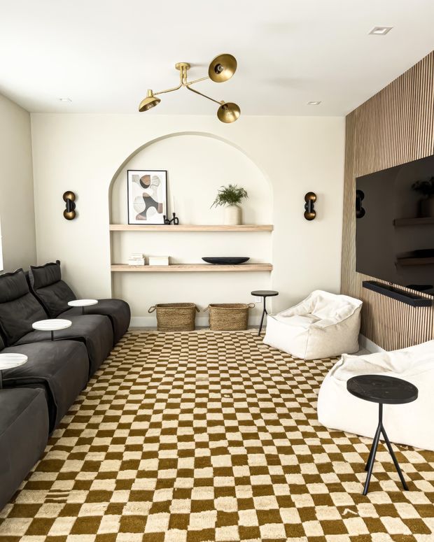 A warm, contemporary living room with a low black sectional sofa, white beanbag-style lounge chairs, round black side tables, and a bold brown-and-cream checkerboard rug. The room features a large arched wall niche with floating timber shelves, sculptural decor, a brass multi-arm ceiling light, brass wall sconces, and a timber slatted media wall surrounding a large television. The palette is neutral with brass and brown accents, and the styling blends modern forms with mid-century-inspired lighting and furniture.