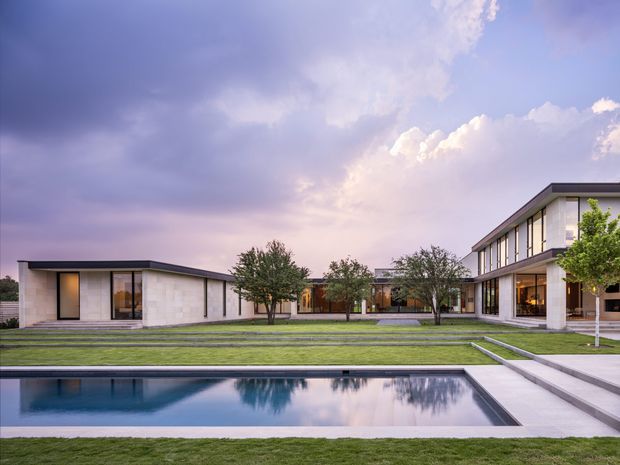 Wide exterior view of a low-slung contemporary residence arranged around a landscaped courtyard and long rectangular swimming pool. The home has crisp horizontal rooflines, extensive floor-to-ceiling glazing, pale masonry or rendered walls, and a restrained geometric composition. A manicured lawn, stepping paths, mature trees, and pool terrace frame the architecture beneath a dramatic pastel evening sky.