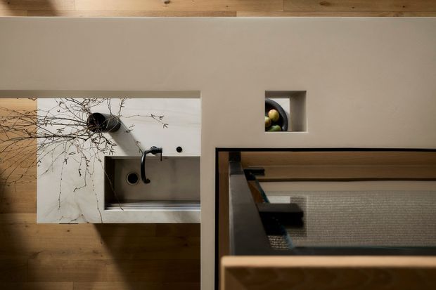 View looking down into a compact contemporary kitchen with warm timber flooring and ceiling, a white marble-look benchtop, stainless-steel sink and tap, and a small recessed display niche. A glazed internal partition or stair opening with timber framing occupies the foreground.