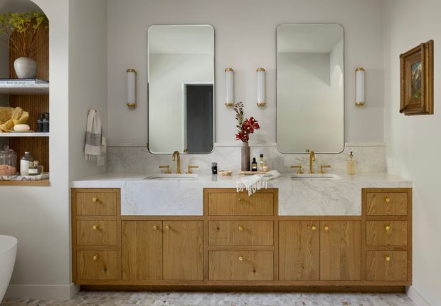A refined double-vanity bathroom with a long timber vanity featuring multiple drawers and cupboards, pale marble-look benchtops and splashback, brushed gold tapware and wall sconces, and two rounded-rectangle mirrors. Open shelving with towels and accessories is visible at left, while a freestanding bath edge appears in the foreground. The room has warm, traditional detailing with a calm neutral palette.