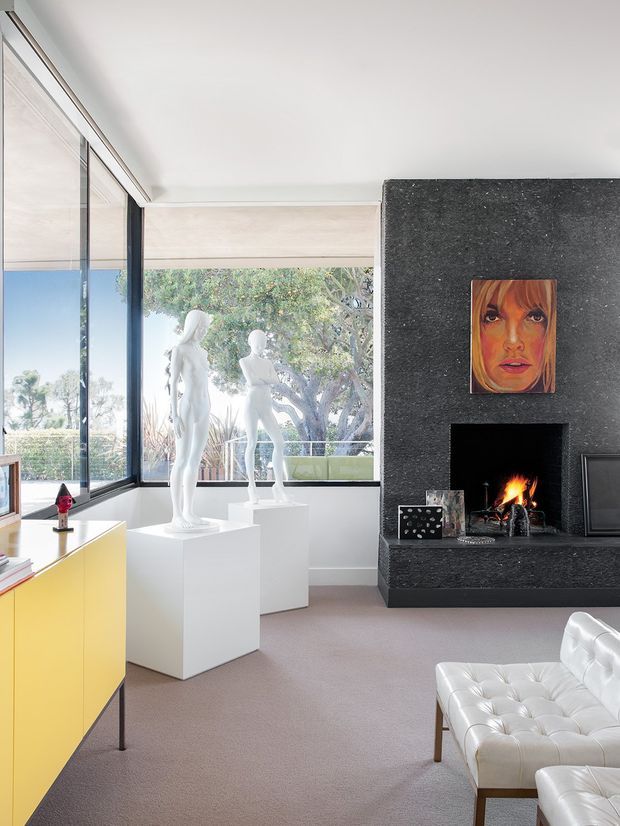 Bright contemporary living room with full-height corner glazing overlooking leafy surroundings. White sculptural figures stand on plinths beside the windows, while a dramatic dark textured stone fireplace wall with a lit fire forms the main feature. A vivid yellow built-in cabinet, white tufted leather seating, artwork, and pale carpeting create a polished gallery-like interior.