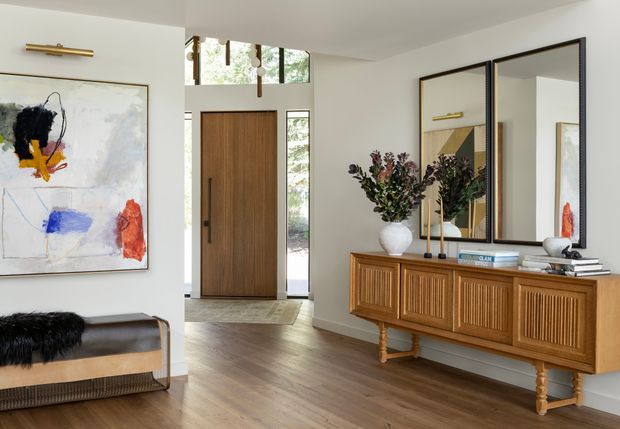 Refined residential entryway with warm timber flooring, a tall natural timber front door, white walls, and high clerestory glazing. A carved timber console with fluted cabinet fronts sits beneath large black-framed mirrors, accompanied by sculptural decor and abstract artwork. The space is bright, warm, and carefully styled with a blend of traditional detailing and contemporary finishes.