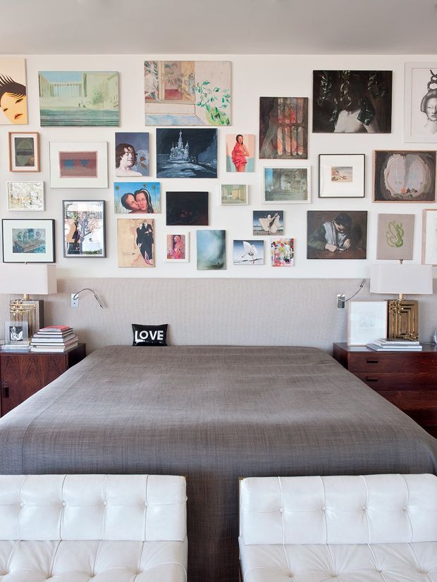 A stylish bedroom with a large upholstered bed covered in grey textured bedding, flanked by dark timber bedside tables with modern lamps. Two white tufted lounge chairs sit at the foot of the bed. The white wall behind the bed is arranged as a dense gallery display of varied framed artwork. The room has a refined, eclectic character with neutral tones and dark timber accents.