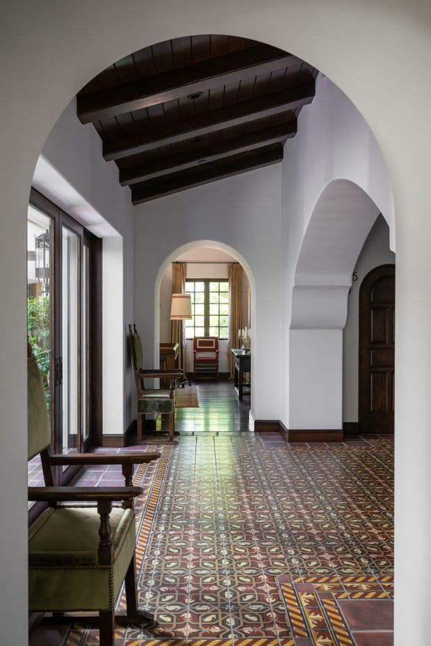A refined interior hallway or entry passage with white plaster walls, multiple rounded arches, dark exposed timber ceiling beams, richly patterned decorative tile flooring, dark timber door and trim, and tall glazed doors along the left side. The passage leads to a small sitting area with traditional upholstered chairs, a lamp, and a window beyond. The composition emphasizes Mediterranean-influenced architectural detailing and a warm, historic character.