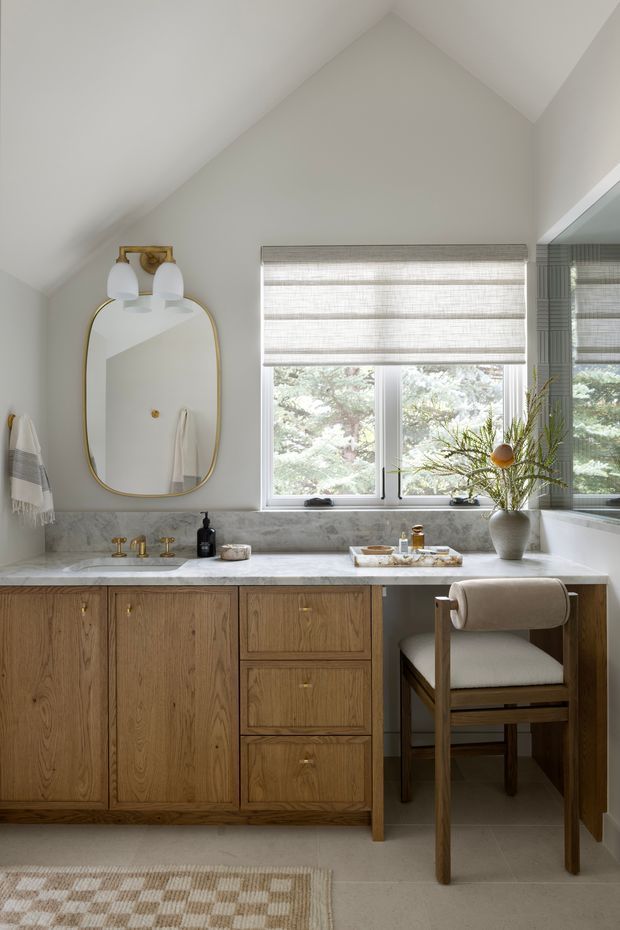 A refined bathroom vanity beneath a steeply sloped ceiling, featuring warm oak cabinetry with brass hardware, a pale stone benchtop and backsplash, twin brass basin fixtures, an arched brass-framed mirror, wall sconces, a wide window with woven roller blind, and a freestanding upholstered vanity chair. The space is softly lit and styled with neutral tones and natural materials.