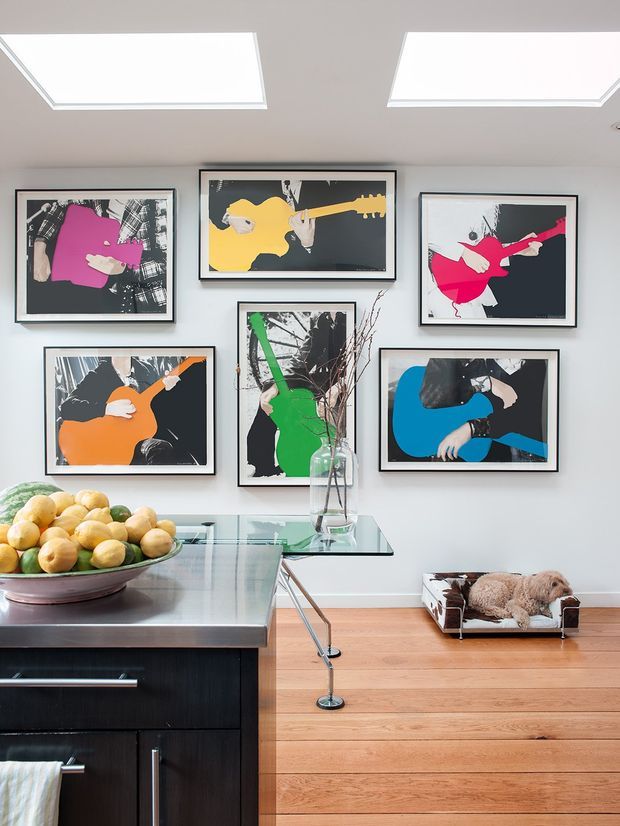 A bright, contemporary kitchen and dining area with a dark kitchen island in the foreground, a glass dining table, light timber flooring, and white walls. The wall is arranged with six colourful framed artworks depicting guitar players. Two rectangular skylights provide strong natural light, while a glass vase with branches and a fruit bowl add decorative elements. A small dog bed is visible on the floor.
