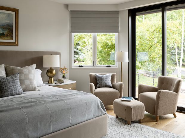 A bright, refined bedroom with a large upholstered bed, layered grey bedding, matching upholstered armchairs and ottoman, bedside table and lamps. Full-height black-framed glazing opens the room visually to a leafy outdoor setting, while a roller blind, pale walls, timber flooring and a neutral patterned rug complete the space.