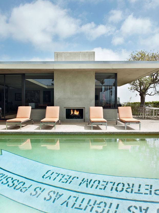 A modern flat-roofed house exterior centered on a pool terrace. The concrete facade features extensive dark-framed glazing, a built-in outdoor fireplace, and a row of peach upholstered loungers facing a pale green swimming pool. A dining area and landscaped tree are visible to the right, beneath a bright blue sky.