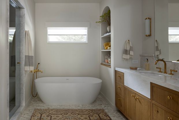 A bright, refined bathroom featuring a freestanding oval bathtub, glass-enclosed shower, double vanity with warm wood cabinetry and a white stone benchtop, brass tapware, a built-in open shelf, and patterned floor tiles. Neutral white walls and natural light create a calm, elegant atmosphere.