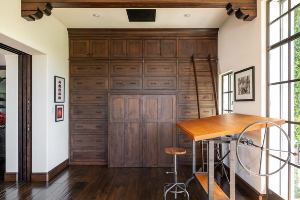 A compact, richly detailed home office or study with a full-height dark timber built-in wall of cabinetry, drawers, and central cupboard doors. A timber-topped drafting or work table with metal supports and stools occupies the foreground beside large black-framed windows. Dark hardwood flooring, exposed dark timber ceiling beams, white walls, and framed artwork create a warm, refined, traditional atmosphere.