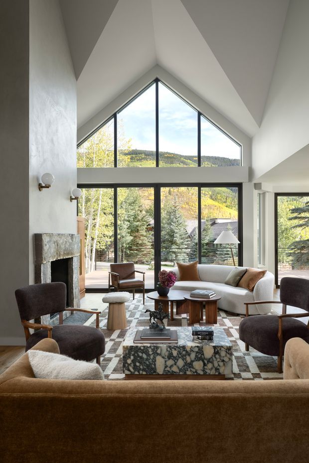 A sophisticated living room with a dramatic vaulted, peaked ceiling and expansive black-framed floor-to-ceiling glazing overlooking a wooded mountainous landscape. The room features a large stone fireplace, a pale upholstered sofa, multiple dark lounge chairs, a brown velvet sofa in the foreground, a sculptural stone coffee table, layered area rugs, and warm contemporary furnishings. The architecture is bright and open, with strong vertical glazing and a high gabled volume.