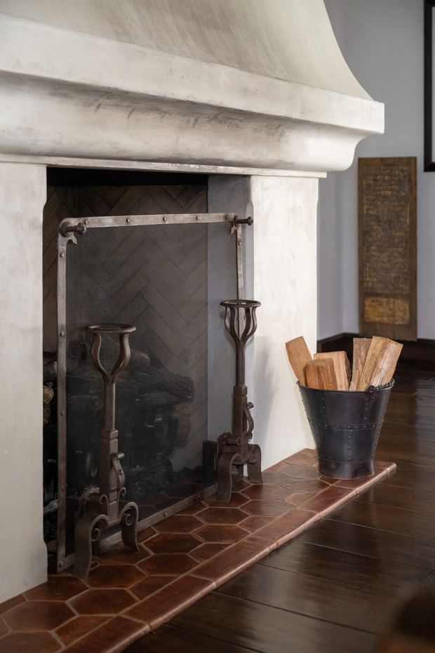 Close-up interior view of a traditional fireplace with a large sculpted white plaster mantel, dark herringbone firebox lining, and ornate aged-metal fire screen and tools. The hearth is finished in reddish-brown irregular terracotta tiles, adjoining dark timber flooring. A black metal firewood bucket and stacked logs sit beside the fireplace, with a partially visible rustic wall panel in the background.