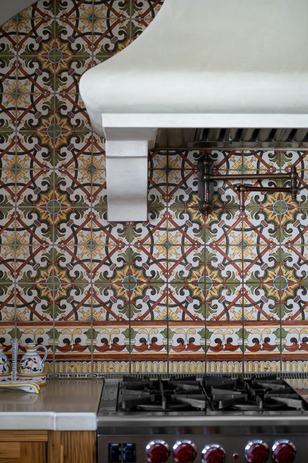 Close-up of a richly patterned decorative tile backsplash in a traditional kitchen. The tiles feature intricate multicolour geometric and floral motifs with a decorative border. A large white sculptural rangehood sits above a stainless-steel gas cooktop and oven, with dark metal pot rails and wooden cabinetry visible below.
