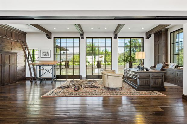A spacious, refined home office with dark timber built-in cabinetry and exposed ceiling beams. Large black-framed grid windows and glazed doors bring in abundant natural light and overlook a landscaped outdoor setting. The room features a substantial timber desk, a central desk or console, a leather armchair, a patterned area rug, and traditional-style furnishings.
