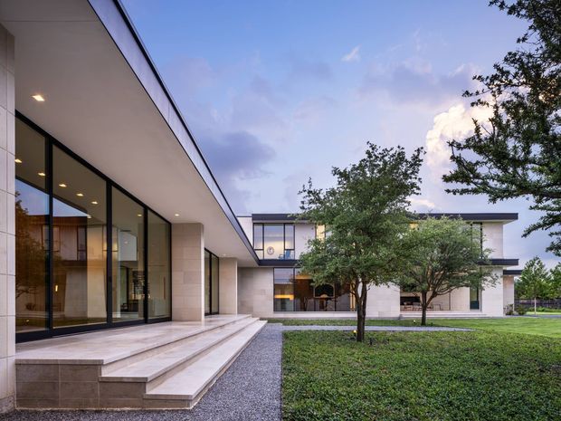 A contemporary modernist house viewed from the landscaped courtyard, with broad low-pitched roof planes, extensive floor-to-ceiling glazing in dark frames, pale stone or rendered wall surfaces, and a stepped terrace entry. The composition features clean horizontal lines, a gravel path, clipped lawn, and mature trees around the building.