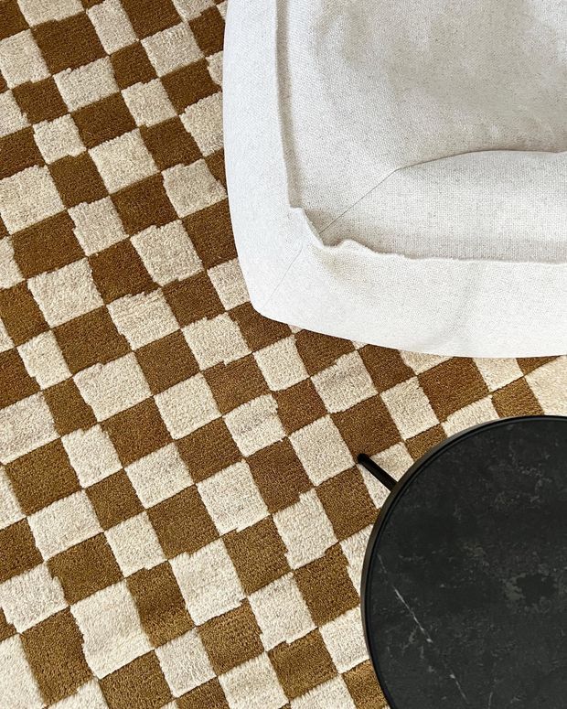 Close-up interior detail showing a brown-and-cream checkerboard textured rug beneath a white upholstered sofa or armchair, with part of a round black side table visible in the foreground.