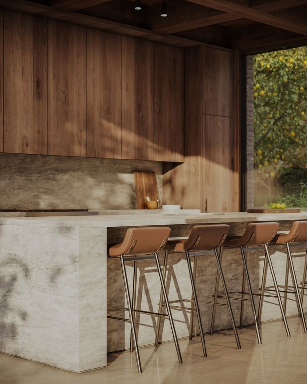 A warm, moody contemporary kitchen centred on a long island with seating for five or more. The space features extensive dark timber wall and ceiling panelling, a pale stone island and benchtop, darker stone backsplash and island sides, slim metal-framed bar stools with brown leather seats, and a large window overlooking greenery.