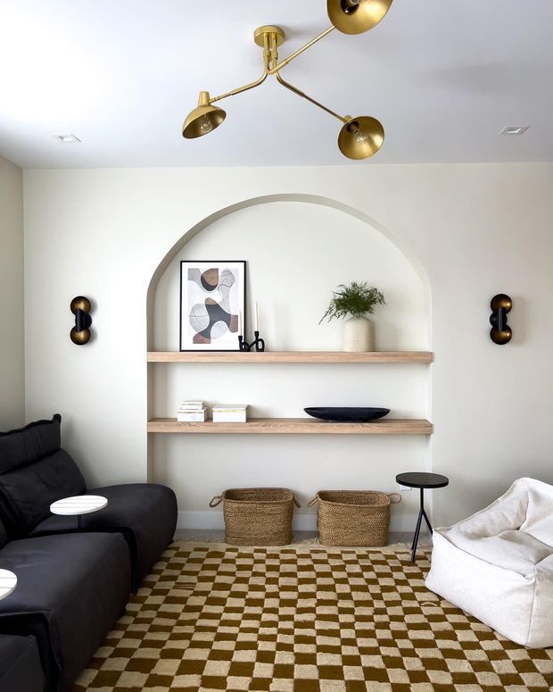 A bright, carefully styled living room with a large arched built-in shelving niche, pale walls, light timber shelves, and sculptural decor. The room includes a dark curved sofa, a white lounge chair, woven baskets, a checkerboard area rug, and striking brass and black wall and ceiling lights.